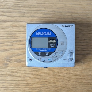 May include: A silver Sharp MD-MT161 portable MiniDisc recorder featuring a circular blue display area with text including MD-MT161, MD PORTABLE RECORDER, DIGITAL RECORDING, and 24bit ATRAC. The device includes various control buttons for playback, recording, and volume.