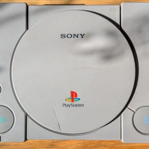 May include: A light grey Sony PlayStation games console featuring a circular disc cover with the Sony logo, a colourful PlayStation emblem, a reset button, a power button with a blue icon, and an eject button with a blue icon on the front panel.