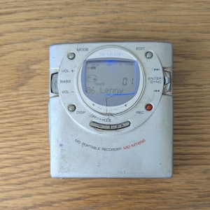 May include: A silver Sharp MD-MT866 portable MiniDisc recorder featuring a circular central display screen with blue backlighting. The device includes various control buttons labelled Mode, Edit, Vol, Bass, Disp, Rec, and Enter/Sync, with the text 06 Lenny visible on the screen.