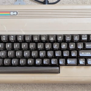 May include: A beige Commodore 64 home computer featuring a QWERTY keyboard with dark brown keys, a power indicator light, and a rainbow logo. The keyboard includes function keys on the right side and a space bar, with text labels on the keys.