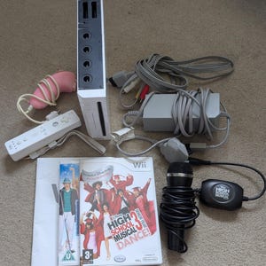 May include: A white Nintendo Wii console bundle including a white Wii remote, a pink Nunchuk, a grey power adapter, AV cables, a black wired microphone with a High School Musical branded adapter, and the High School Musical 3 Senior Year Dance game.