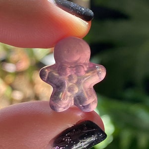 May include: A small, translucent pink gingerbread man figurine. The figurine is held between two fingers, with a blurred background of greenery and sunlight. The gingerbread man has a classic shape with arms and legs outstretched.