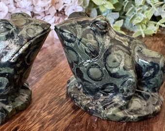 Puede incluir: Dos figuras decorativas de rana. Las ranas están hechas de una piedra moteada verde y negra. Las figuras de rana están sobre una superficie de madera. Las ranas se miran. El fondo incluye flores blancas y vegetación.