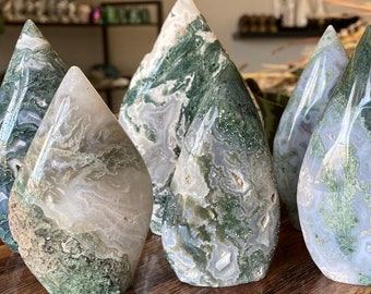 May include: A selection of teardrop-shaped, polished moss agate stones. The stones feature a mix of white, grey, and green hues, with intricate patterns. They are arranged on a wooden surface, showcasing their natural beauty.
