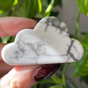 May include: A white cloud-shaped stone with gray veining is held in a hand. The polished stone has a smooth surface and a cloudy pattern. The background features green foliage. This decorative stone is a unique home decor item.