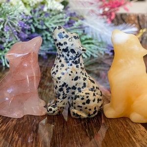 May include: Four carved stone wolf figurines in shades of green, pink, speckled, and yellow. Each wolf is in a howling pose, with their heads tilted upwards. The figurines are displayed on a wooden surface.