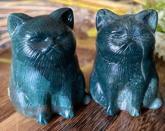 May include: Three dark green cat figurines are displayed on a wooden surface. The cats are carved with detailed features, including closed eyes and a slightly grumpy expression. The figurines are a deep teal color, and the background includes dried flowers.