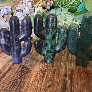 Könnte beinhalten: Vier geschnitzte Kaktusskulpturen aus Stein in Grün-, Lila- und Schwarztönen werden auf einer Holzoberfläche präsentiert. Die Kakteen sind in einer Reihe angeordnet, jeder mit einer einzigartigen Farbe und Muster, die eine natürliche, erdige Ästhetik zeigen.