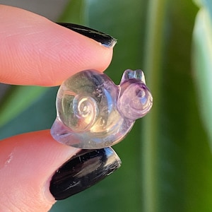 May include: A small, translucent snail figurine carved from a blend of purple and yellow crystal. The snail's shell features a spiral design, and the body is detailed with tiny antennae. The figurine is held between fingers against a blurred green background.
