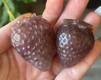 May include: Two carved strawberry-shaped crystals in shades of purple and brown. The crystals have a textured surface, mimicking the appearance of a real strawberry. The crystals are held in a hand, with a blurred green plant in the background.