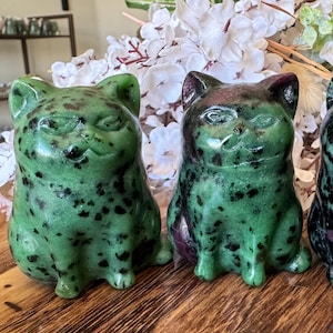 May include: Three carved stone cat figurines. The cats are green with black speckles and a hint of red. They are sitting upright with a slightly smug expression. The figurines are displayed on a wooden surface.