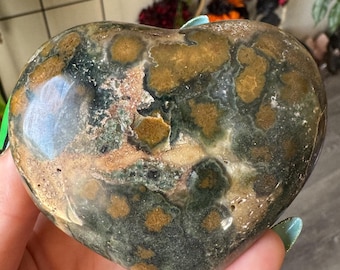 May include: A heart-shaped stone with a mottled pattern of green, brown, and tan. The polished surface reflects light, highlighting the natural variations in colour and texture. This decorative stone is a unique home decor item.
