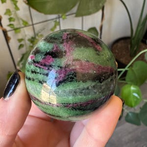 Pode incluir: Uma pedra esférica polida com uma mistura vibrante de inclusões minerais verde-escuras, pretas e rosa magenta. A superfície é lisa e reflexiva, exibindo variações de cores naturais e padrões por todo o objeto redondo.