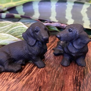 May include: Two small, black dachshund figurines are displayed on a wooden surface. The figurines are detailed, with visible ears, legs, and facial features. The background includes green and white foliage.