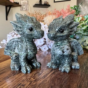 Op de afbeelding: Twee gedetailleerde, groene stenen drakenbeeldjes met grote ogen en vleugeldetaills. De draken zitten op een houten oppervlak, met een achtergrond van bloemen en groen. De beeldjes zijn ongeveer 10 cm hoog.