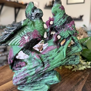 Op de afbeelding: Een gebeeldhouwde stenen sculptuur van twee vogels die op een tak zitten. De vogels en de tak zijn groen met rode en zwarte aderen. De sculptuur wordt tentoongesteld op een houten oppervlak.