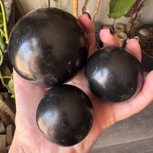 May include: Three polished black spheres of varying sizes are held in a hand. The spheres have a smooth, reflective surface, and the lighting creates highlights. The largest sphere is at the top, with two smaller ones below.