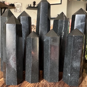 Pode incluir: Uma coleção de obeliscos de cristal preto polido com pontas pontiagudas. Os cristais variam em altura e estão dispostos sobre uma superfície de madeira. Os cristais têm uma superfície refletora e são exibidos em ambientes internos.