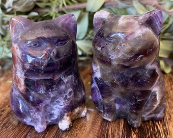 May include: Two carved amethyst cat figurines. The cats are seated, with detailed facial features and a polished finish. The stone displays shades of purple, brown, and white. The figurines are displayed on a wooden surface.