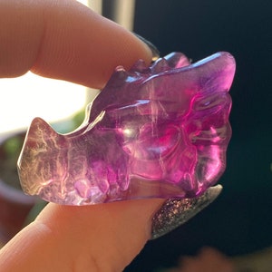 May include: A translucent, purple and white fluorite carving in the shape of a dragon's head. The gemstone is held in a person's hand, with the dragon's head facing left. The carving has detailed features and a polished surface.