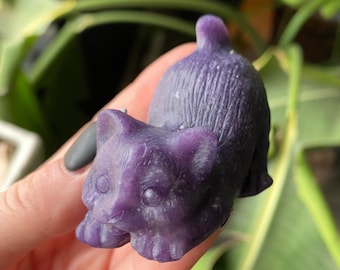 May include: A carved purple cat figurine. The cat is depicted in a playful pose, with its front paws extended and its tail raised. The figurine is made of a smooth, polished material and has detailed features.