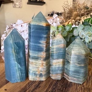 Pode incluir: Quatro torres de cristal de calcita azul de vários tamanhos são exibidas em uma superfície de madeira. Os cristais têm uma aparência em camadas com listras azuis, brancas e bege. Os cristais são polidos e têm pontas pontiagudas. O fundo inclui vegetação e itens decorativos.