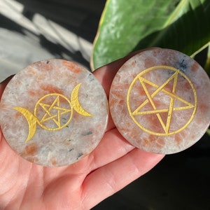 Può includere: Due sottobicchieri rotondi in pietra solare con simboli dorati. Uno presenta un design a tripla luna e pentagramma, l'altro un pentagramma. I sottobicchieri hanno un aspetto maculato arancione e bianco.