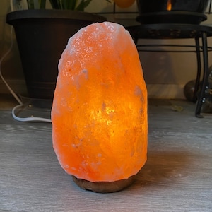 Puede incluir: Una lámpara de sal del Himalaya de color naranja brillante. La lámpara tiene una superficie rugosa y texturizada y se asienta sobre una pequeña base de madera redonda. Emite una luz cálida y acogedora, creando un ambiente acogedor. Es un artículo de decoración del hogar.