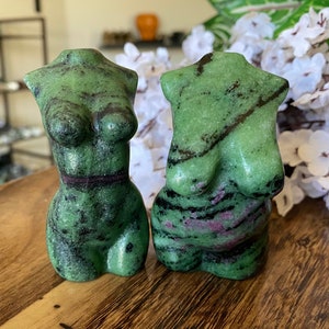 May include: Two green and red stone torso sculptures. The sculptures are carved in the shape of a female torso. The stone has a green base color with black and red veining throughout. The sculptures are displayed on a wooden surface.