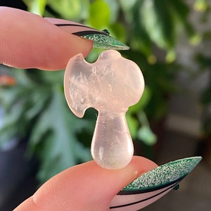 May include: A small, light pink crystal axe figurine. The axe head is rounded, and the handle is a smooth, elongated shape. The crystal is translucent, and the axe is held between two fingers against a blurred green background.