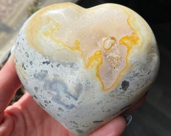 May include: A heart-shaped stone with a mix of colors. The top is a creamy beige with yellow and orange accents, while the sides are a speckled gray and white. The stone has a smooth, polished surface and a natural, organic appearance.