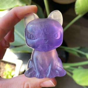 May include: A translucent purple cat figurine with pointed ears and an angry expression. The cat is sitting, with its paws in front. The figurine is held in front of a background of green leaves.