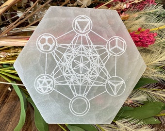 May include: A hexagonal, off-white selenite crystal plate with a white Metatron's Cube design etched on its surface. The plate is surrounded by dried flowers and greenery, set against a wooden background. The crystal measures approximately 6 inches across.