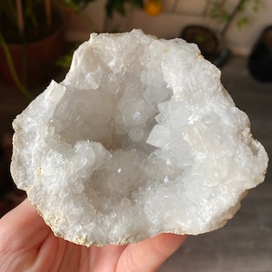 Pode incluir: Geodo de quartzo branco, mostrando um interior cristalino dentro de um exterior áspero bege. O geodo está aberto, revelando as brilhantes formações de cristais pontiagudos no interior. Uma bela pedra natural.
