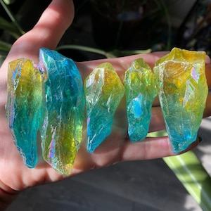 Puede incluir: Cinco cristales iridiscentes con un degradado de colores de amarillo a azul. Los cristales tienen una superficie rugosa y texturizada y se sostienen en una mano. Los cristales son de diferentes tamaños y formas, captando la luz.