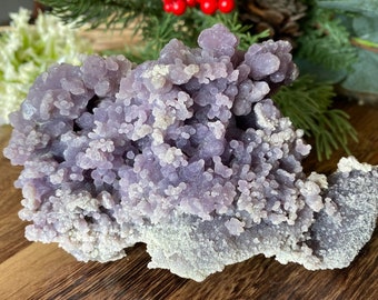 May include: A light purple botryoidal chalcedony cluster, a type of quartz, with a bubbly, grape-like texture. The stone has a light grey base and sits on a wooden surface. Decorative greenery and red berries are in the background.