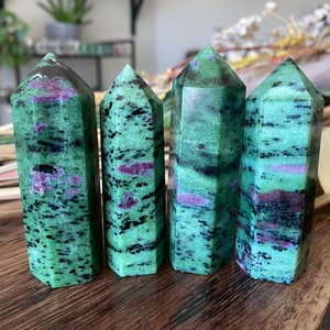 May include: Four green and red ruby zoisite crystal towers with hexagonal shapes. The crystals have a mix of green, black, and red hues. The crystals are standing on a wooden surface.