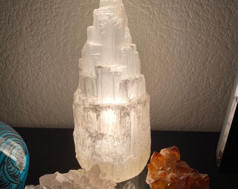 Könnte beinhalten: Eine leuchtende, weiße Selenitlampe mit einem geschichteten, strukturierten Design. Die Lampe wird von innen beleuchtet und wirft ein weiches Licht. Zwei Kristallcluster, einer klar und einer orange, befinden sich vor der Lampe.