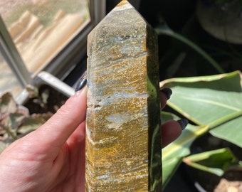 May include: A polished, rectangular crystal obelisk with a pointed top. The stone features bands of gold, brown, and gray hues. The obelisk is held in a hand, with a window and plants in the background. A decorative item.