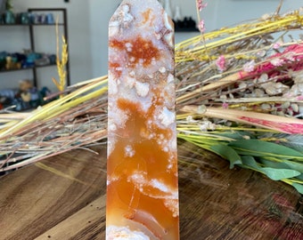 May include: A rectangular crystal tower with a pointed top. The stone is a mix of orange, white, and brown. The crystal is on a wooden surface, with dried flowers in the background. The crystal is approximately 15 cm tall.