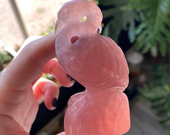 Puede incluir: Una figura de búho tallada en piedra preciosa rosa. El búho está en posición sentada con un agujero para el ojo. La piedra preciosa tiene una calidad translúcida, permitiendo el paso de la luz. El búho se sostiene en una mano.
