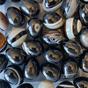 Puede incluir: Una colección de piedras de ágata pulidas, negras y marrones. Las piedras tienen una superficie lisa y brillante con diferentes patrones de rayas y remolinos marrones y blancos. Las piedras tienen forma ovalada y parecen haber sido pulidas o formadas naturalmente.