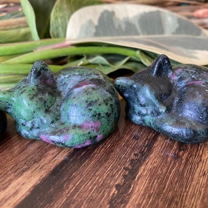 May include: Two small, sleeping cat figurines carved from stone. One is a deep green with red and black flecks, the other is a darker, almost black, version. The cats are curled up, with their tails wrapped around their bodies.