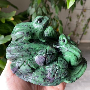 Może przedstawiać: Rzeźba z kamienia przedstawiająca dwie zielone żaby na liściach lilii wodnych. Żaby mają czarne oczy i czarne plamki. Liście lilii wodnych są zielone i fioletowe. Rzeźba jest trzymana w dłoni osoby.