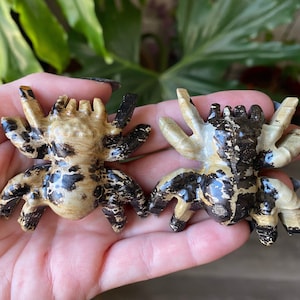 May include: Two carved stone spider figurines. The spiders are tan with dark brown and black markings. The legs are outstretched, and the bodies are detailed. The figurines are displayed against a blurred green background.