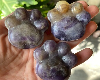 May include: Three amethyst paw-shaped carvings. The carvings are a mix of purple, lavender, and tan colors. The paw prints are smooth and polished, with rounded pads and toes. The carvings are held in a hand.