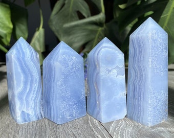 May include: Four polished blue lace agate crystal towers standing upright. Each stone features a distinct light blue colour with white, wavy, banded patterns and a smooth, semi-translucent texture. The points are faceted into a hexagonal shape.