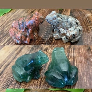 May include: A collection of carved stone frog figurines in various colors. The frogs are in shades of red, gray, green, blue, and pink. Each frog is detailed with visible legs, eyes, and a textured surface. The figurines are displayed on a wooden surface.