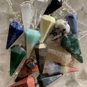 May include: A collection of gemstone pendulums in various colors, including blue, green, black, and clear. Each pendulum is faceted and suspended from a silver chain. The pendulums are arranged on a light-colored surface.