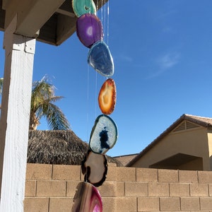May include: A hanging wind chime featuring colorful, polished agate slices in shades of teal, purple, blue, orange, and pink. The agate stones are suspended from a wooden ring, creating a decorative outdoor accent. The wind chime hangs in front of a blue sky.